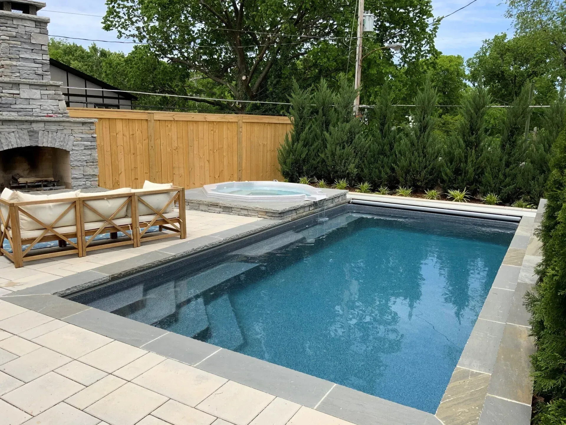 Backyard pool with spa, fireplace, and seating area. Blue water, gray stone surround, and wooden fence.