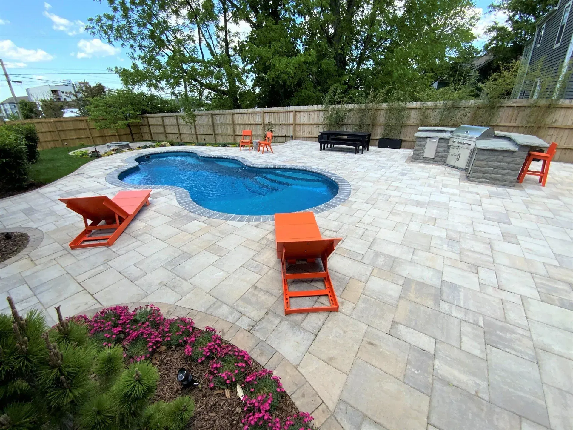 Backyard with a blue pool, stone patio, orange lounge chairs, and an outdoor kitchen area.