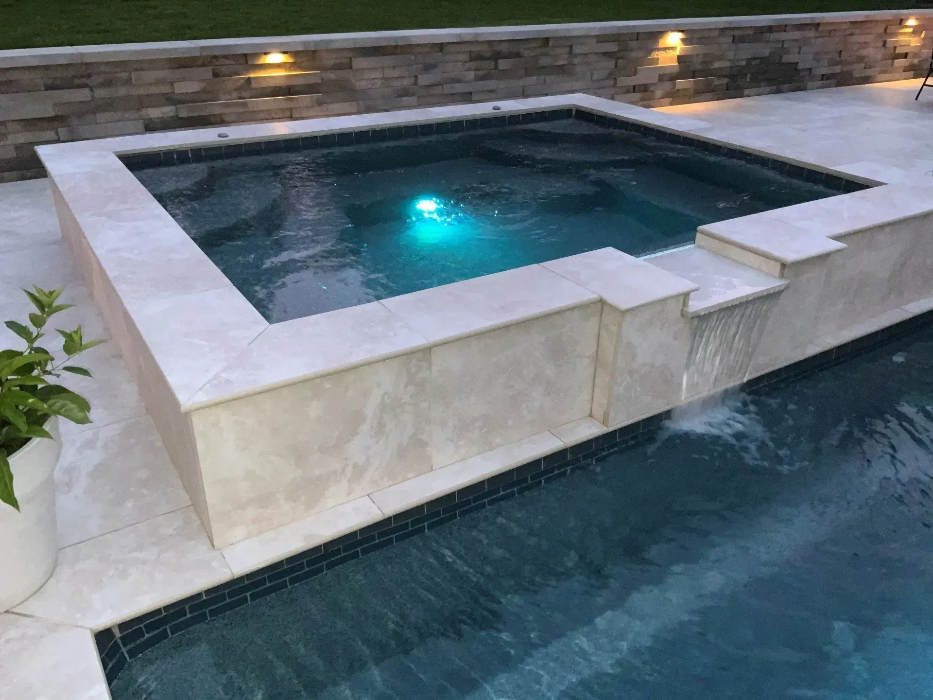 A square spa with waterfall feature flows into a dark blue swimming pool; beige stone surrounds the spa and pool.