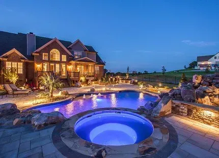 Night view of a luxurious backyard with a pool and hot tub, illuminated lights, and a large house.
