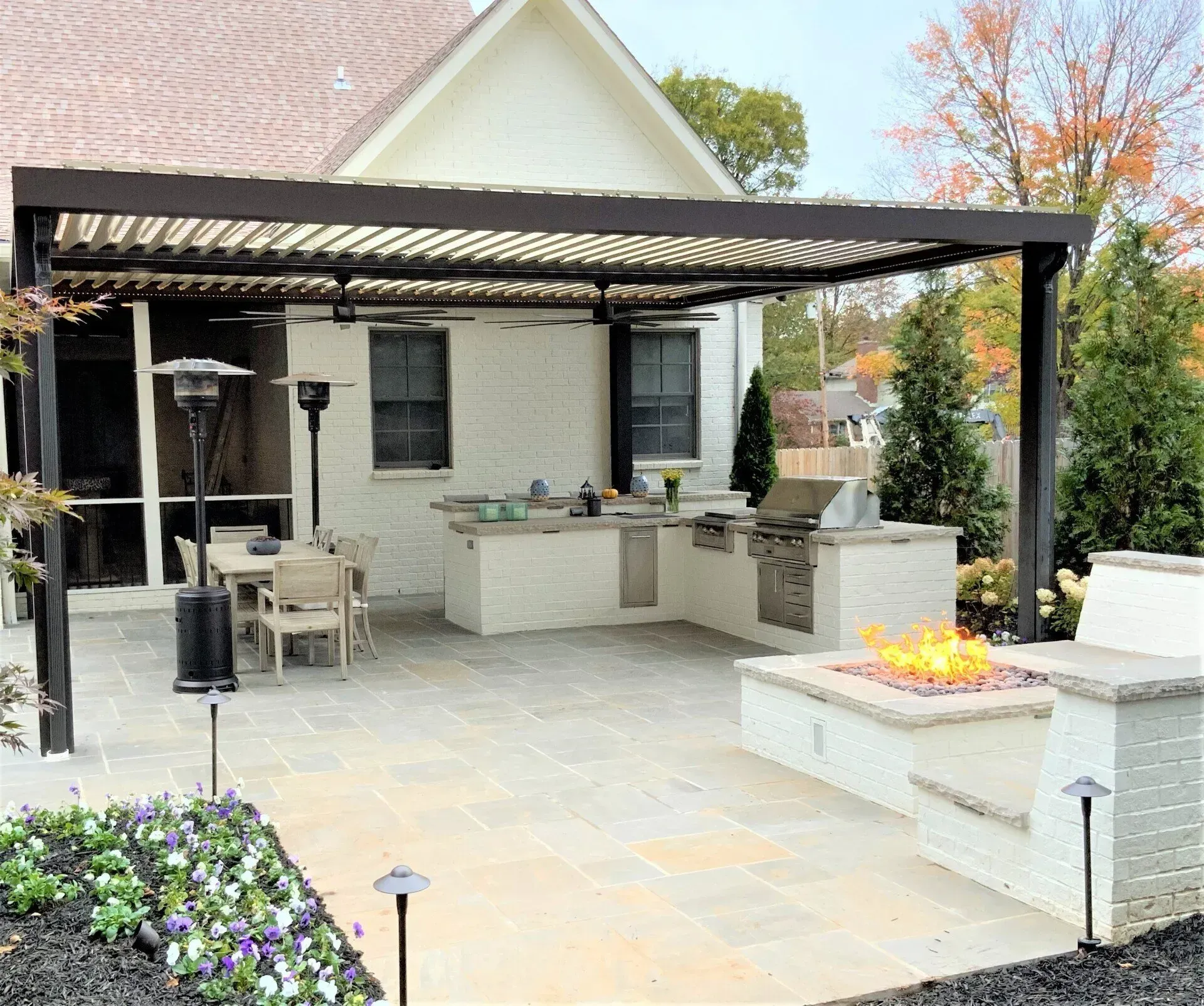 Outdoor patio with kitchen, grill, fire pit, and pergola structure. Beige patio stones and white brick building.