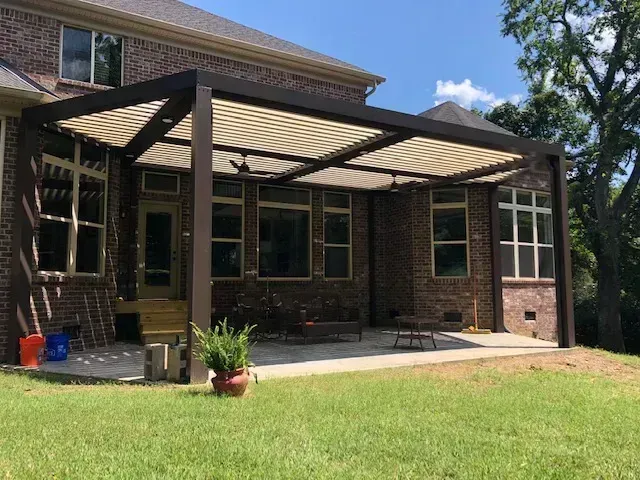 A brown pergola shades a brick patio. Beige canopy, brown supports. Green lawn.