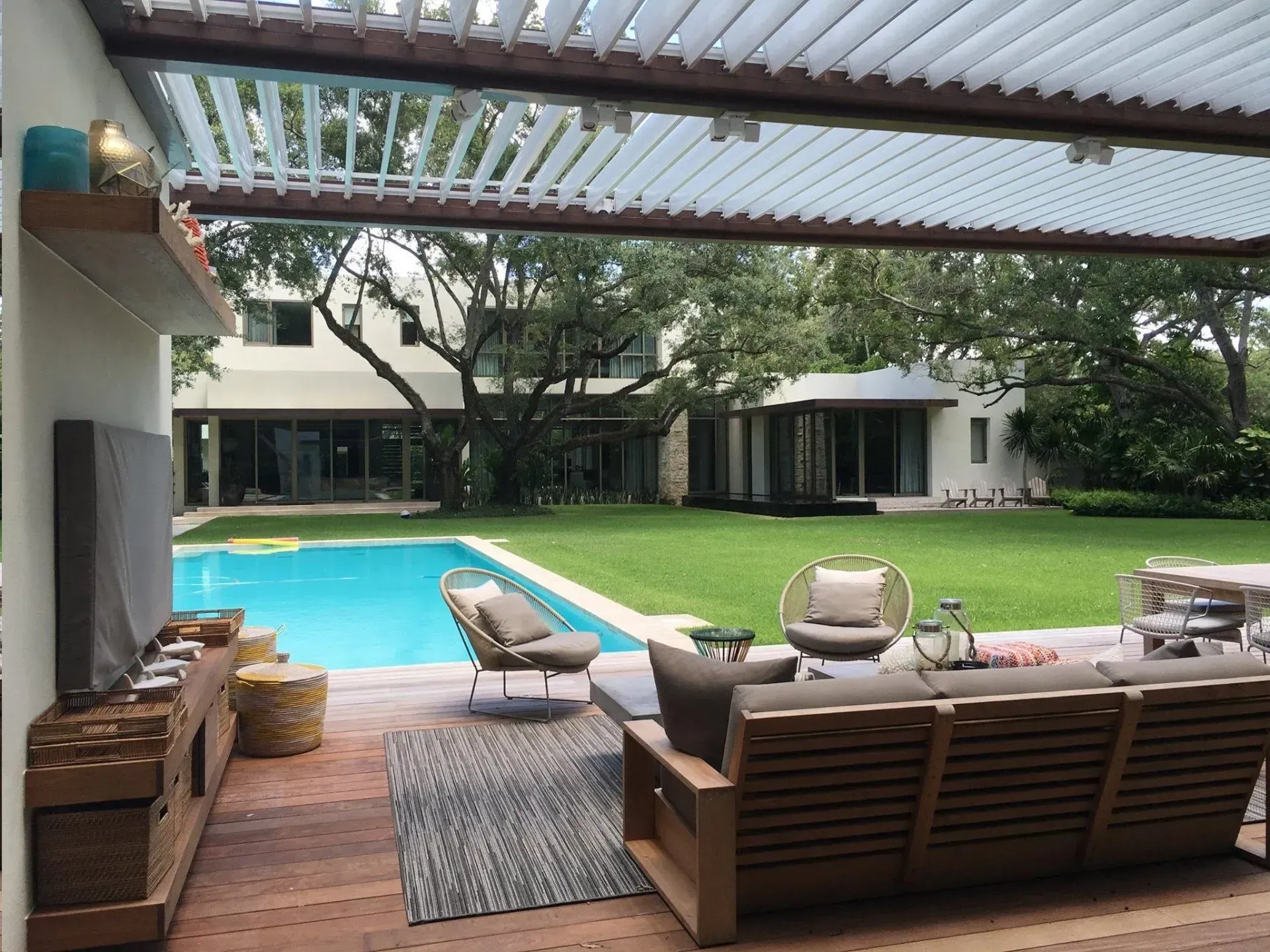 Patio with pool, lawn, and modern house. Wooden furniture, rug, and TV under a pergola.