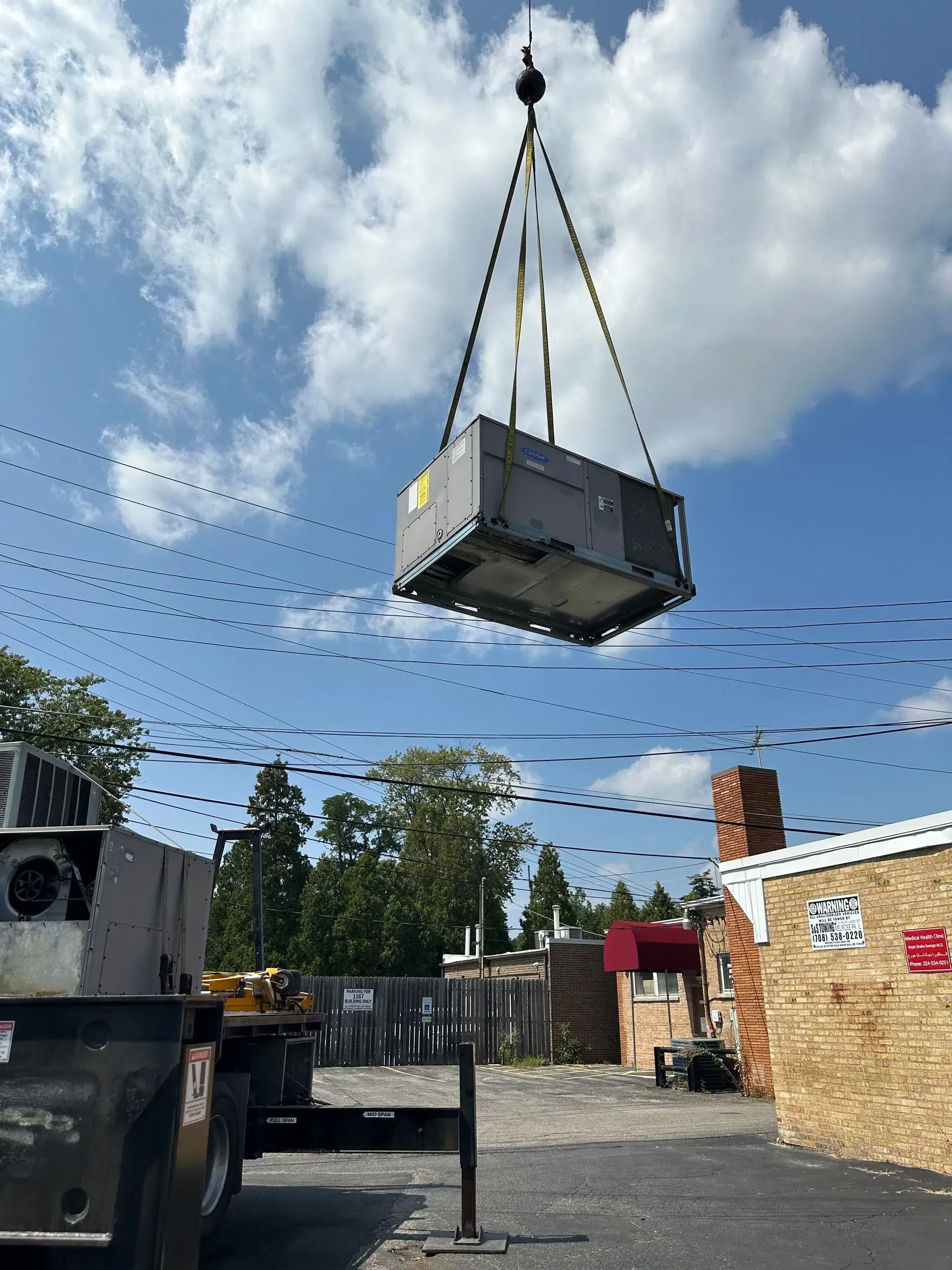 A large HVAC unit is lifted by a crane in front of a building on a sunny day.