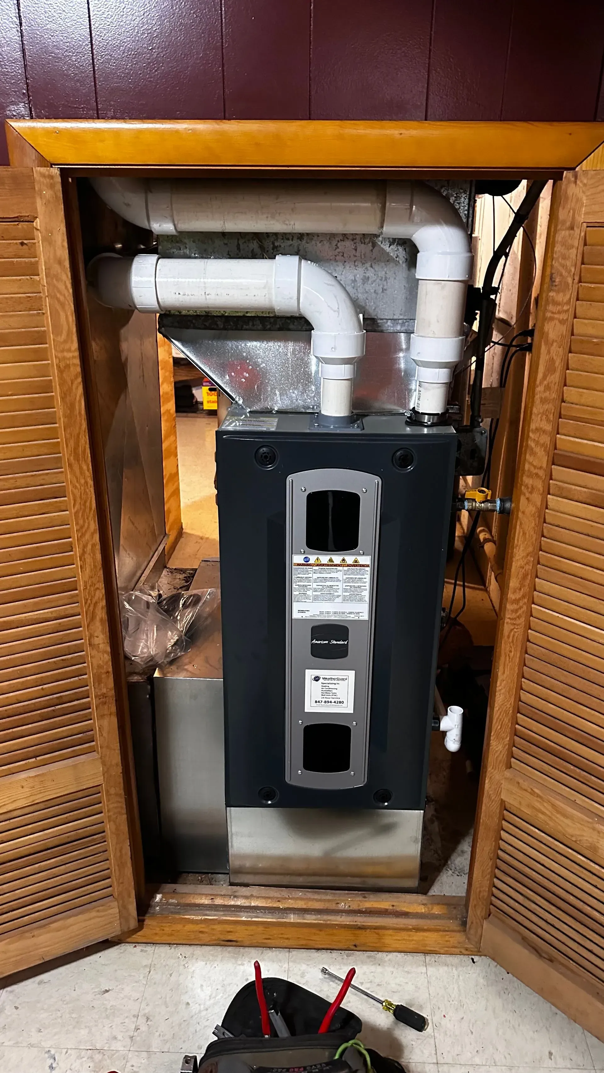 Furnace inside a wooden cabinet, with white pipes and vent; tools in foreground.