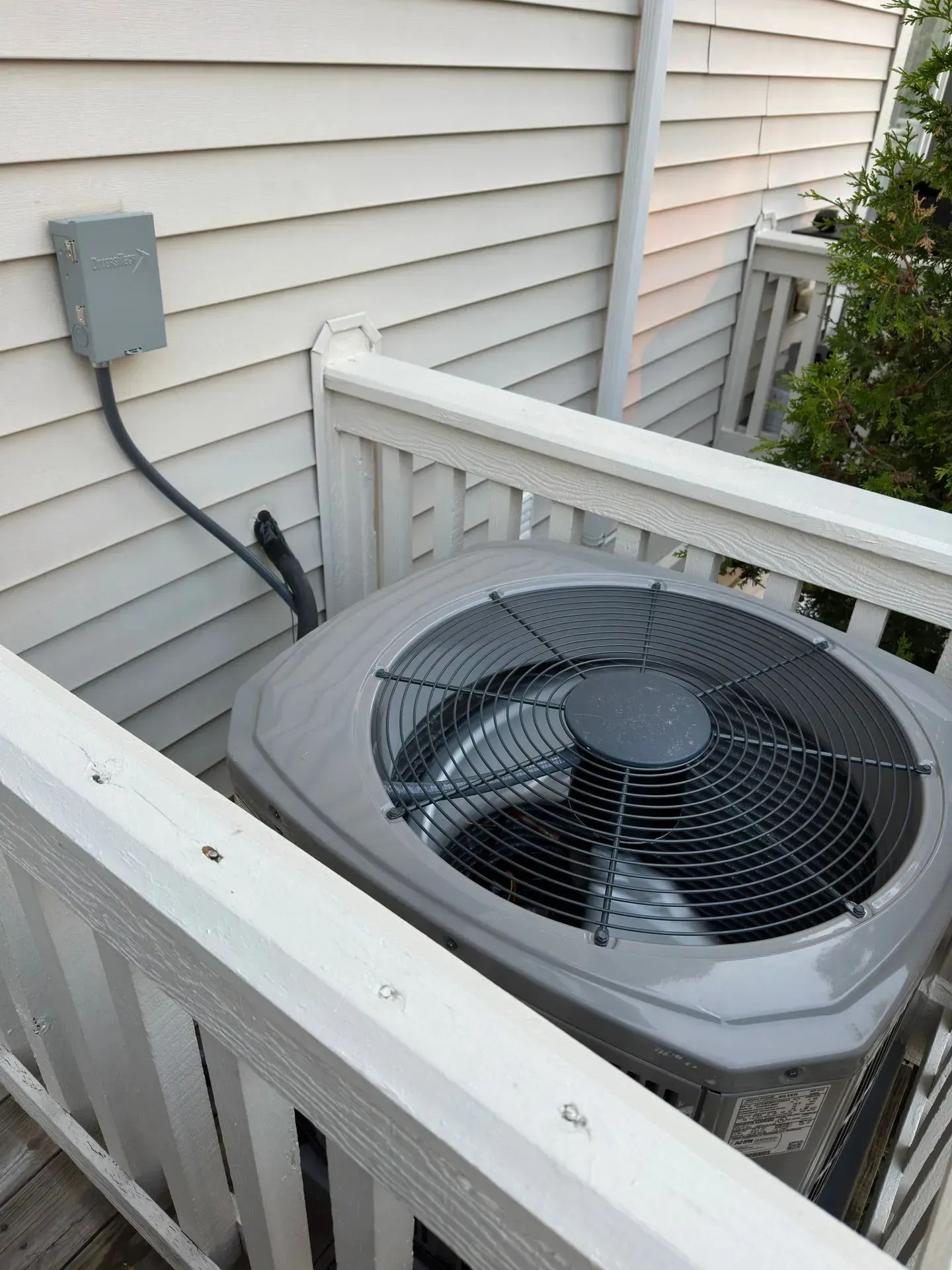 Air conditioning unit on a wooden deck next to a house. Gray unit with black fan.