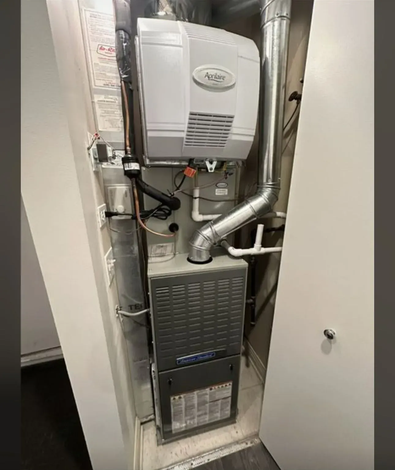 Furnace and humidifier unit inside a white utility closet. Grey metal components with silver ducting and water lines.