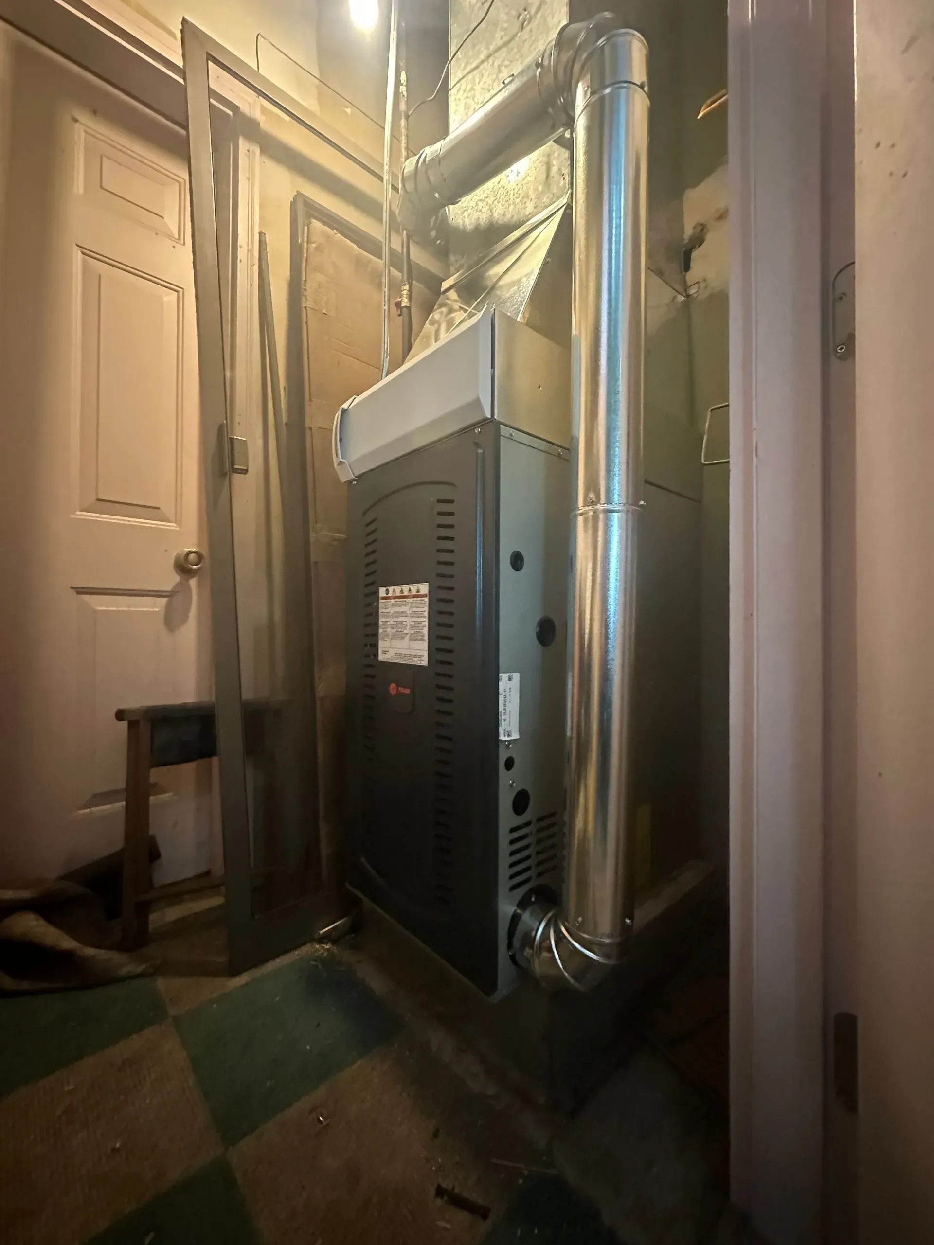 Furnace in a basement. Silver vent pipes, light-colored walls, checkered floor, and a closed door.