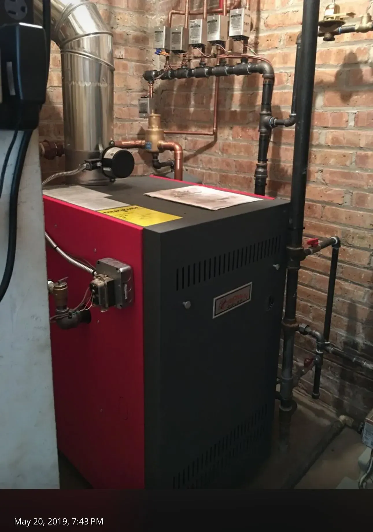Red and black boiler in a basement, with pipes and brick wall visible.