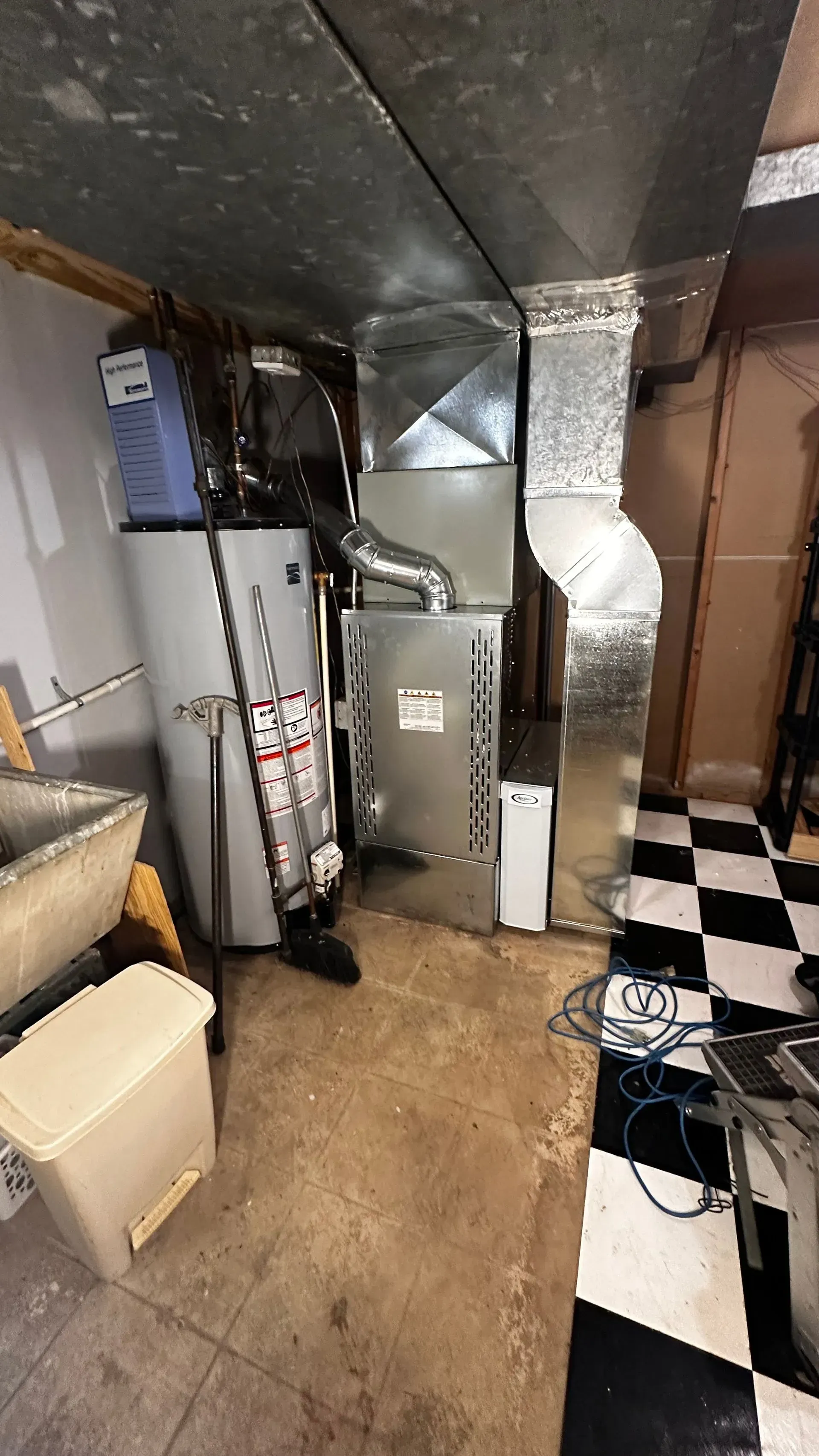 Furnace and water heater in basement, ductwork. Concrete floor with checkered tile.