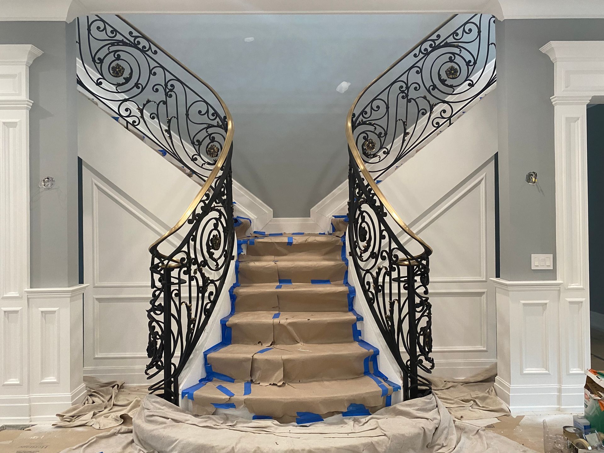 Staircase under renovation with blue tape, covered steps, and ornate black wrought-iron railings