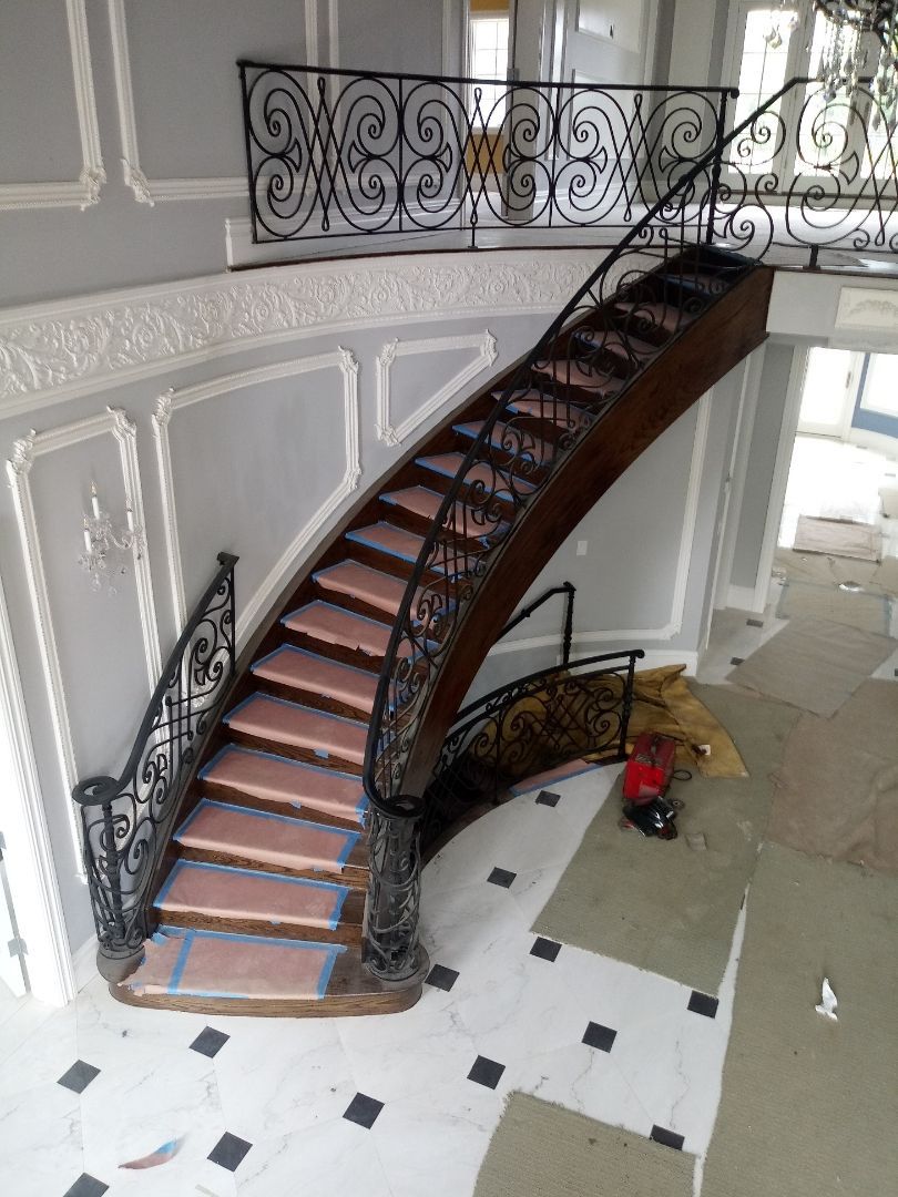 Curved wooden staircase with ornate black railing in a bright, white foyer; red vacuum on the floor below.