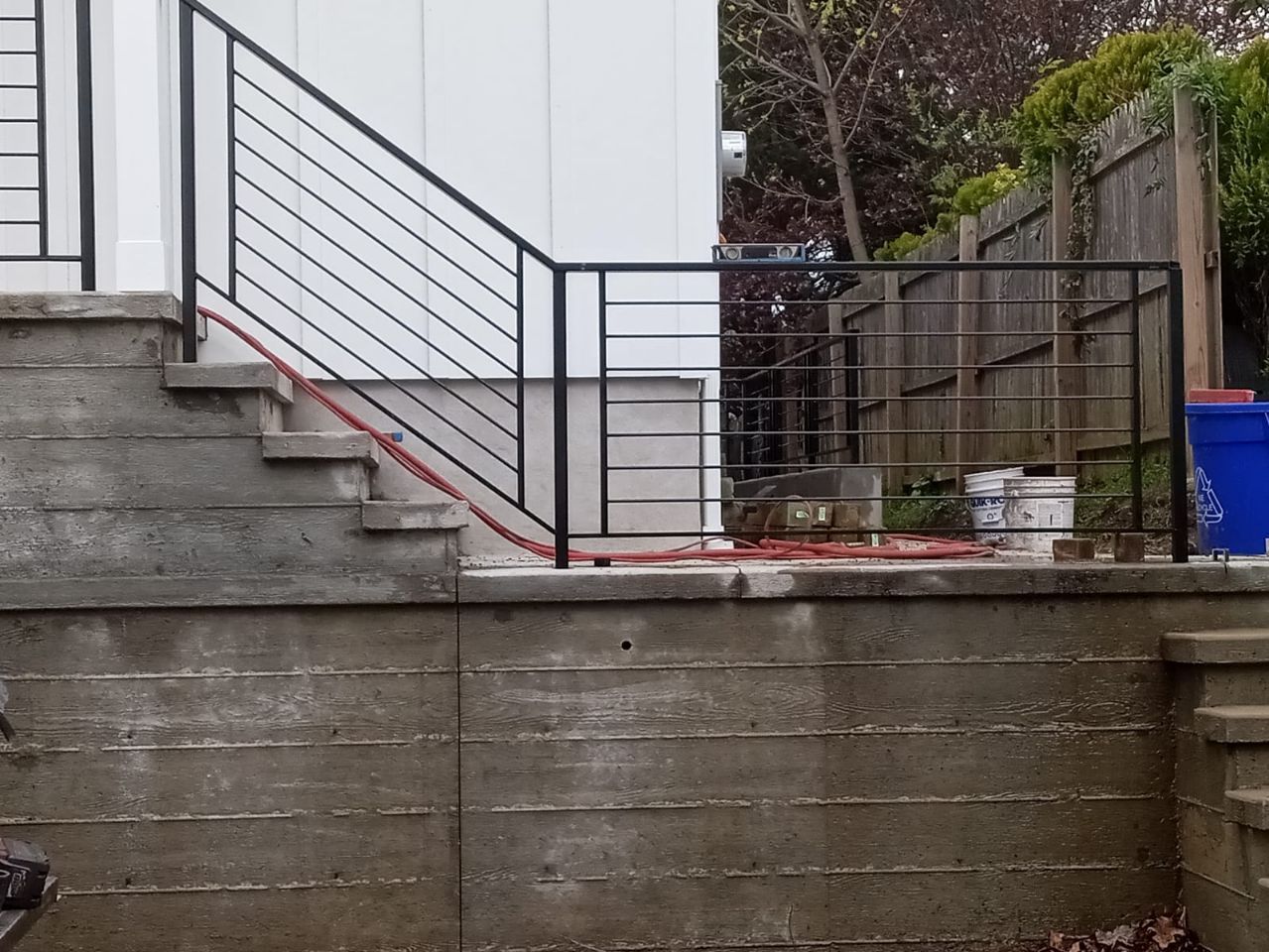 Concrete backyard steps with black metal railing beside a white house and retaining wall