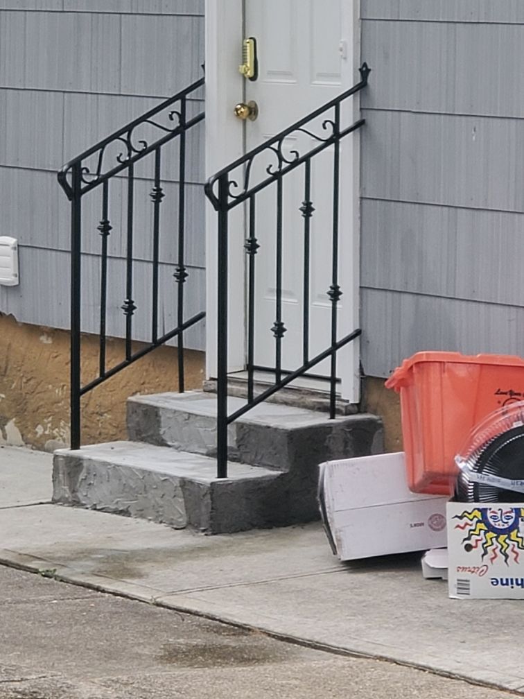Front steps with black metal railings leading to a white door, with trash bins and boxes beside the house.