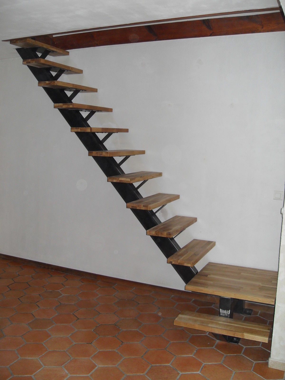 Steep wooden staircase with open treads along a white wall, leading to a loft or upper level.