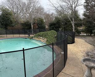 A swimming pool in a backyard surrounded by a black mesh safety fence with trees in the background.