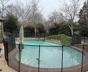 An oval swimming pool with a stone rock feature, surrounded by a black mesh safety fence in a backyard setting.