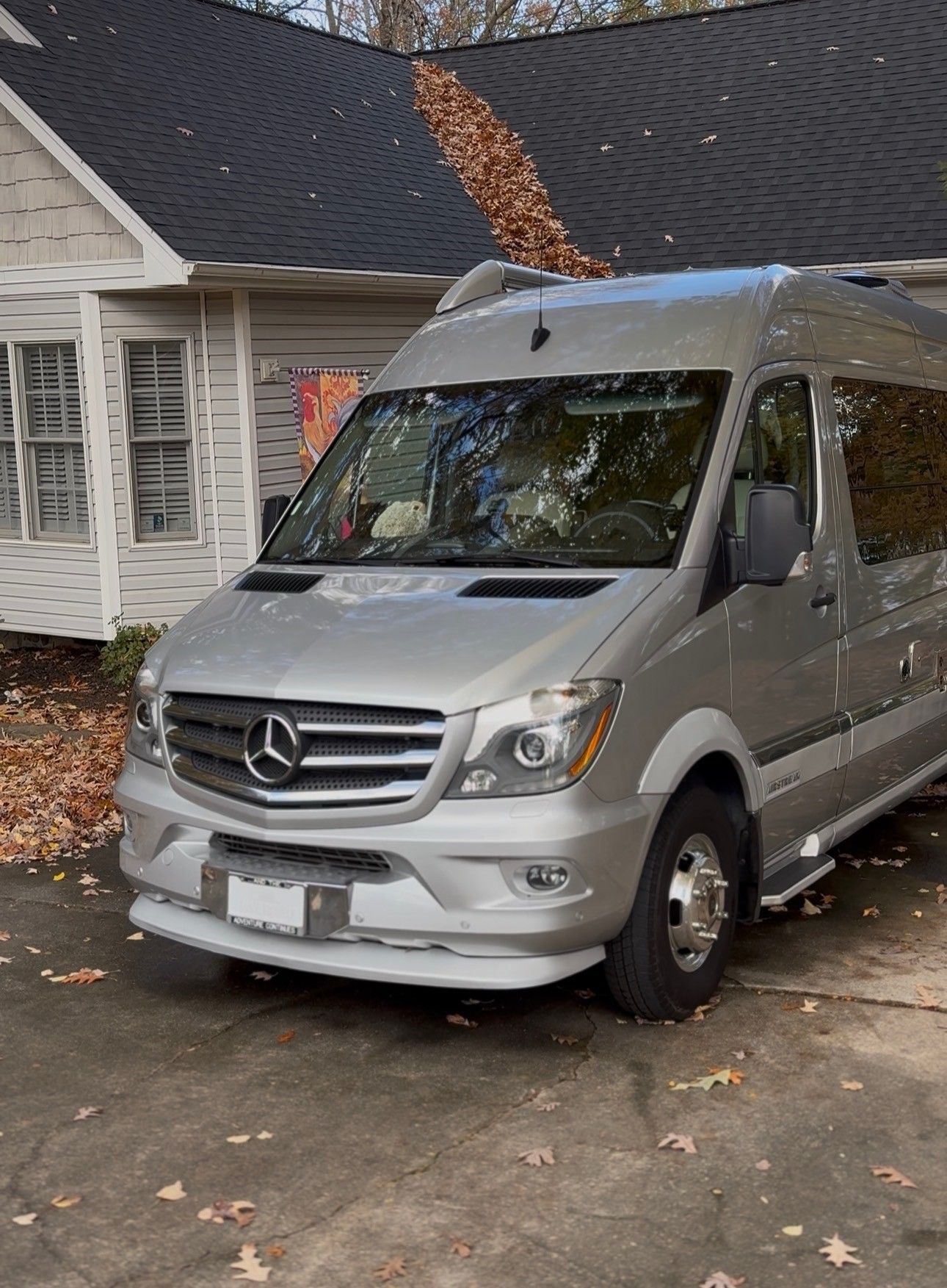 Silver Mercedes-Benz Sprinter van parked in front of a house with fall leaves on the roof.