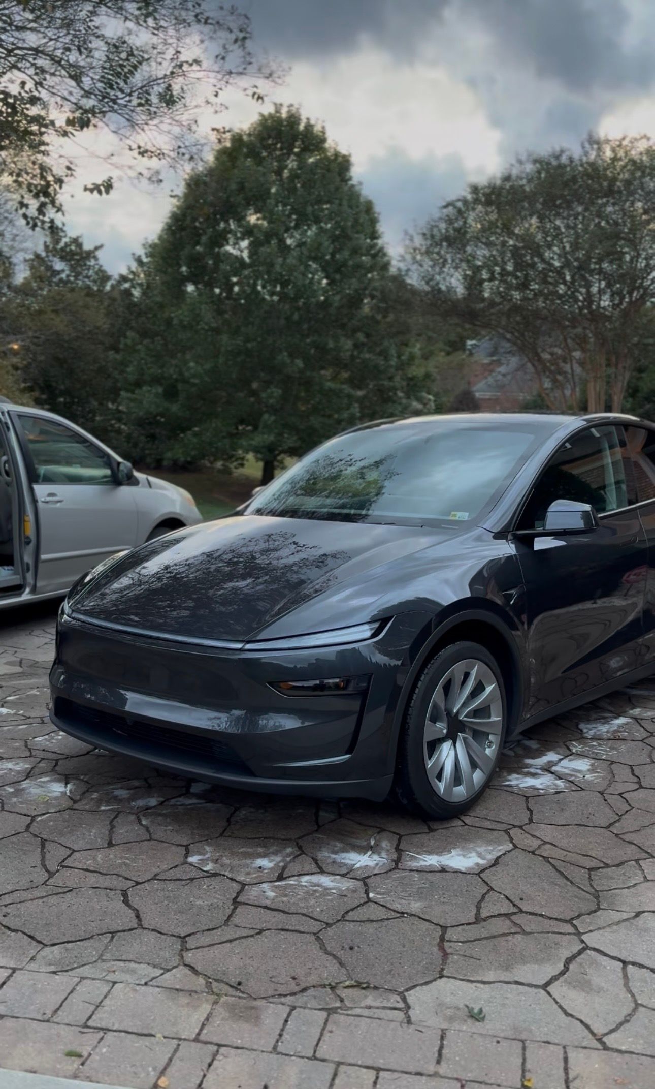 Black Tesla Model Y parked on a stone driveway, next to a silver van, with trees in the background.