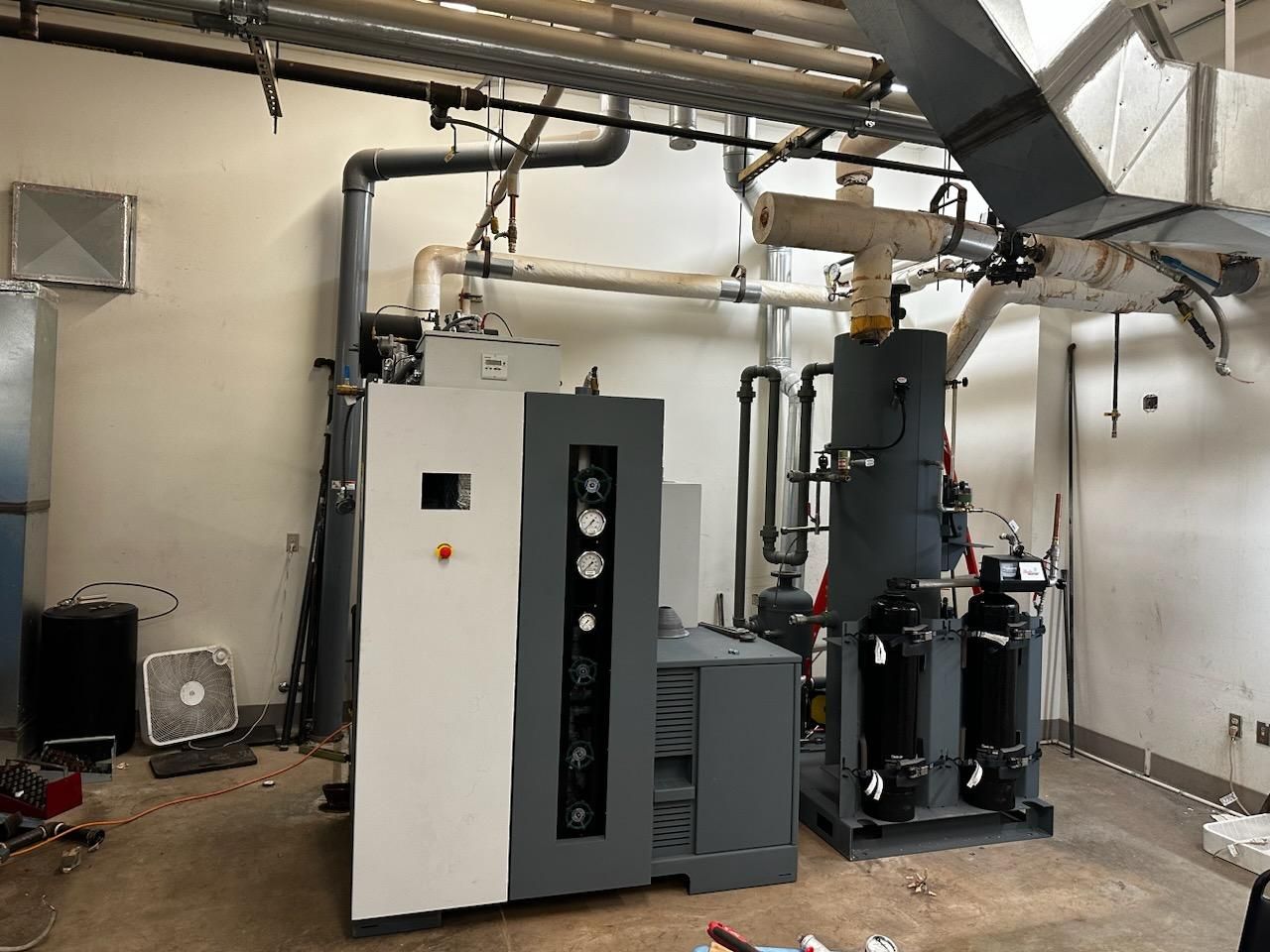 Industrial equipment in a mechanical room, including a large, gray machine with a control panel, pipes, and ventilation.