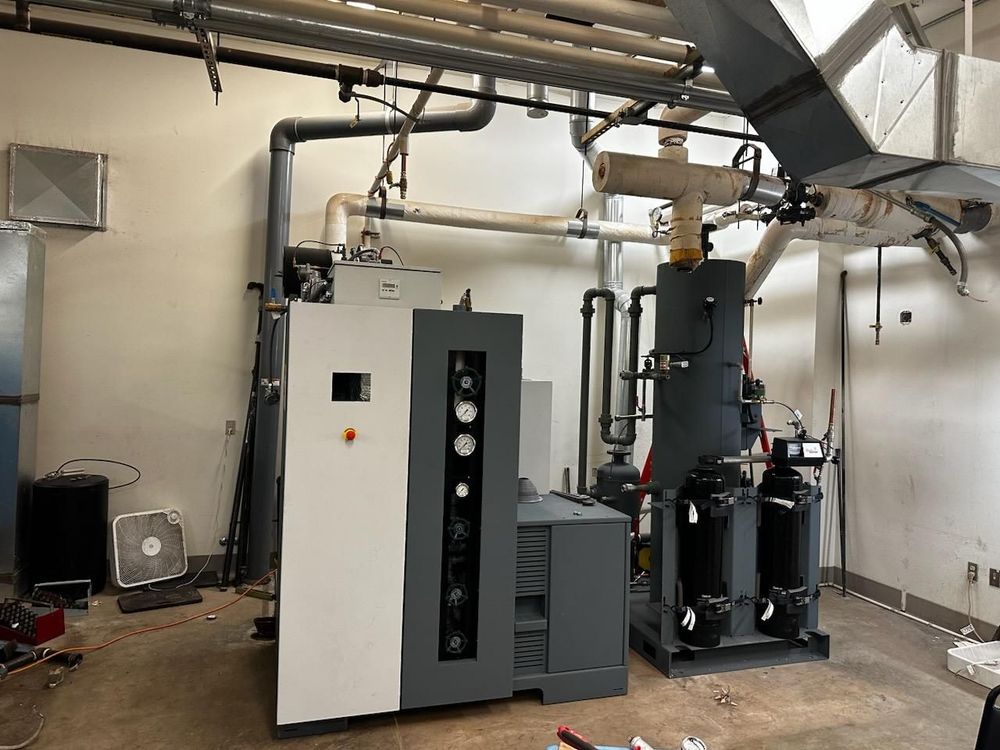 Industrial equipment in a mechanical room, including a large, gray machine with a control panel, pipes, and ventilation.