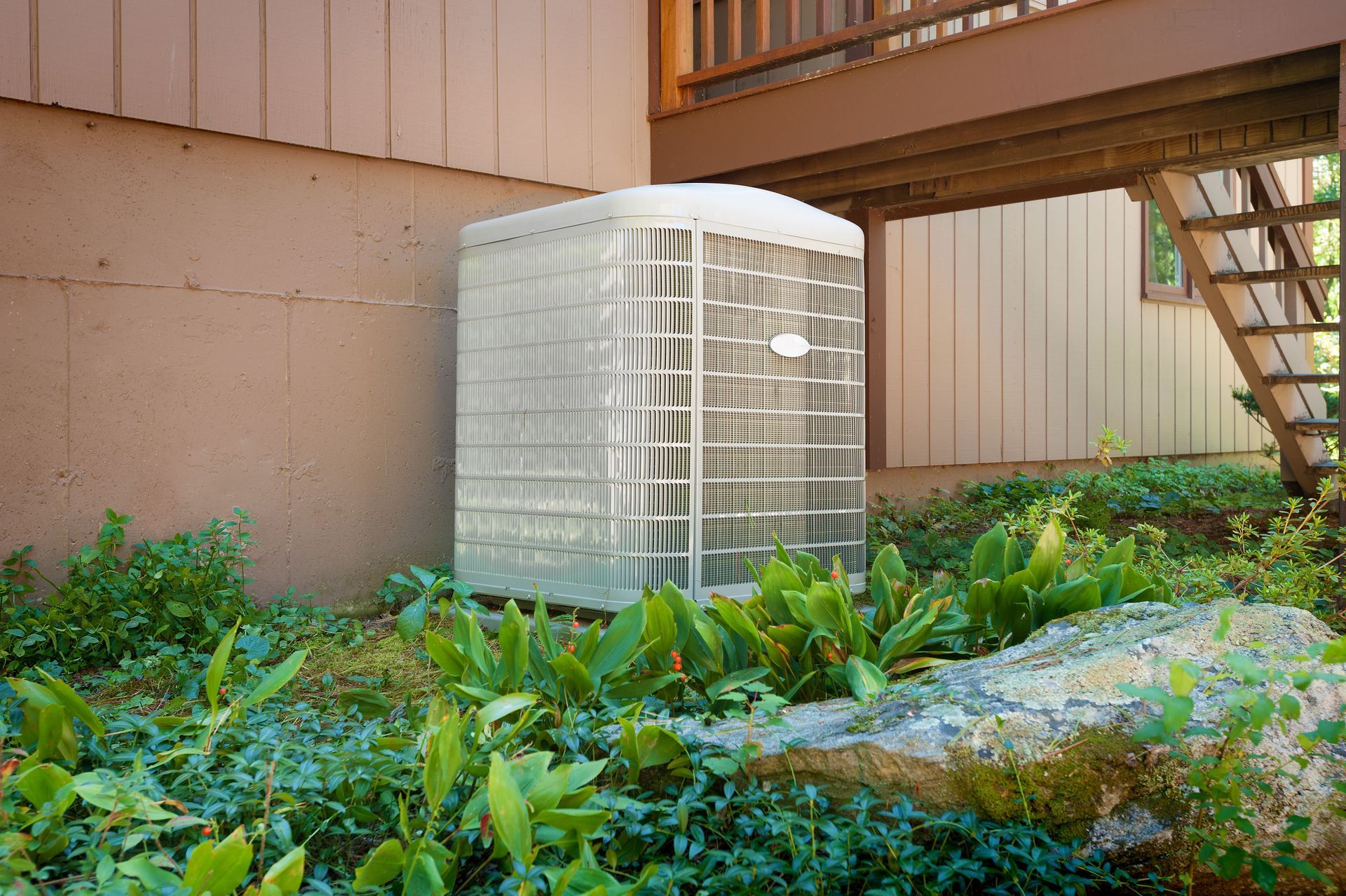 An air conditioning unit sits amongst greenery beside a building with a wooden deck and stairs.