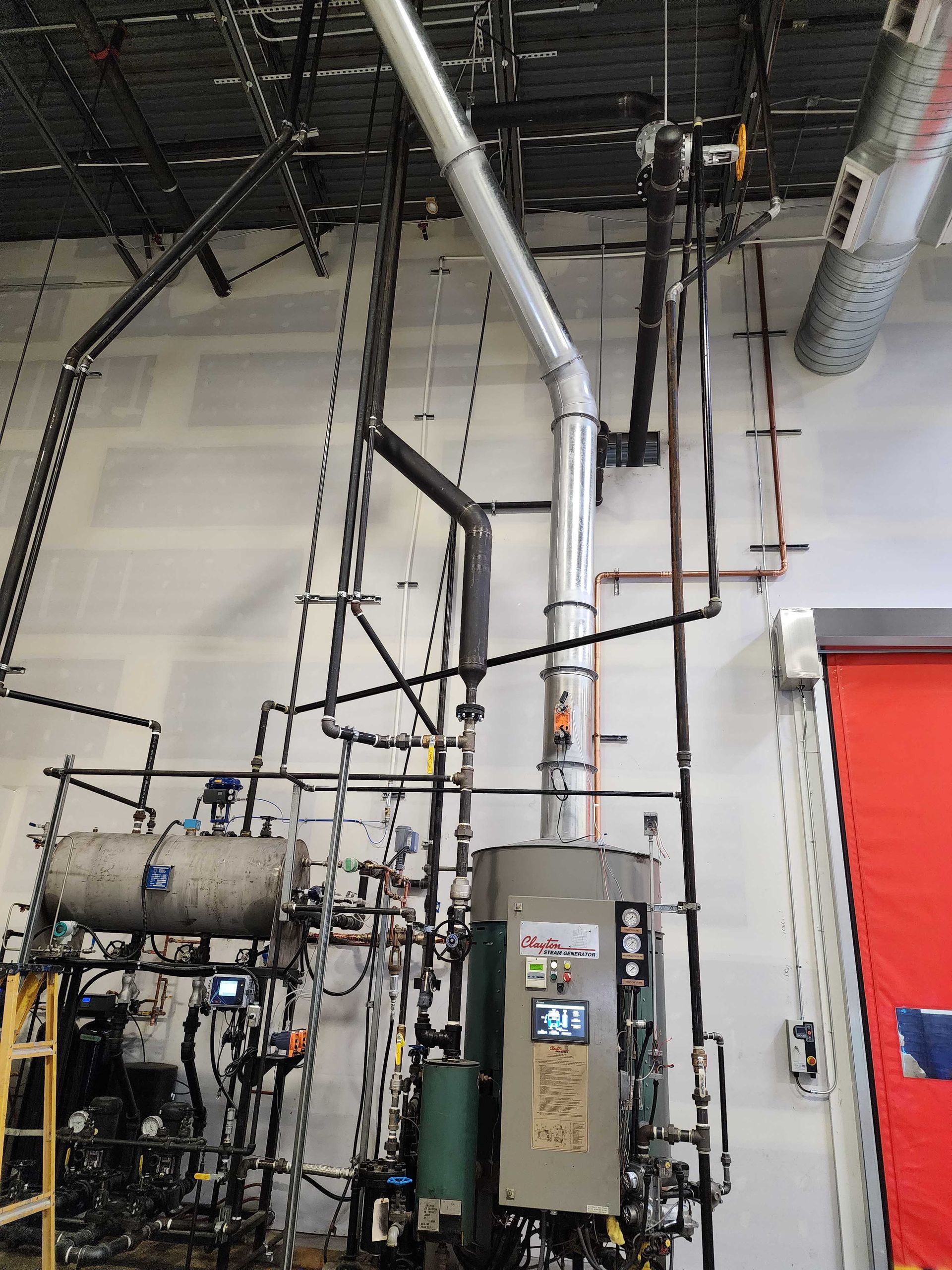Industrial equipment with pipes and a control panel against a white wall; the view includes a red door and ceiling fixtures.