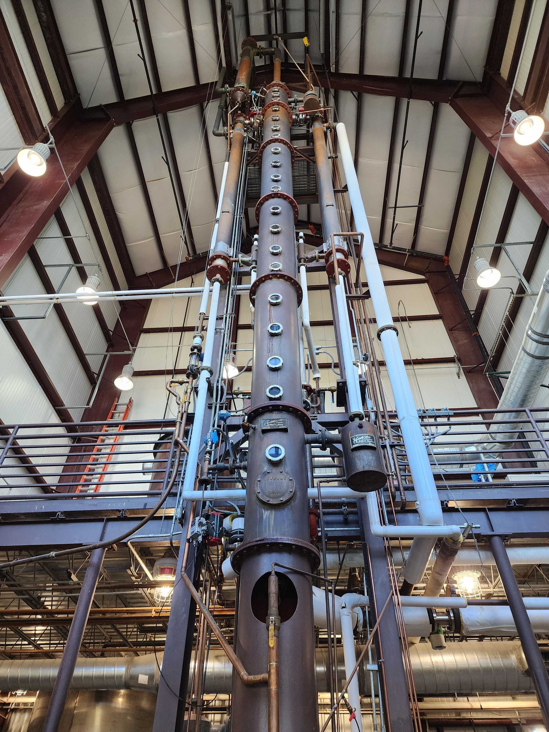 A tall, industrial still for distillation, inside a building with a metal frame roof. Copper pipes and gauges are attached to the column.
