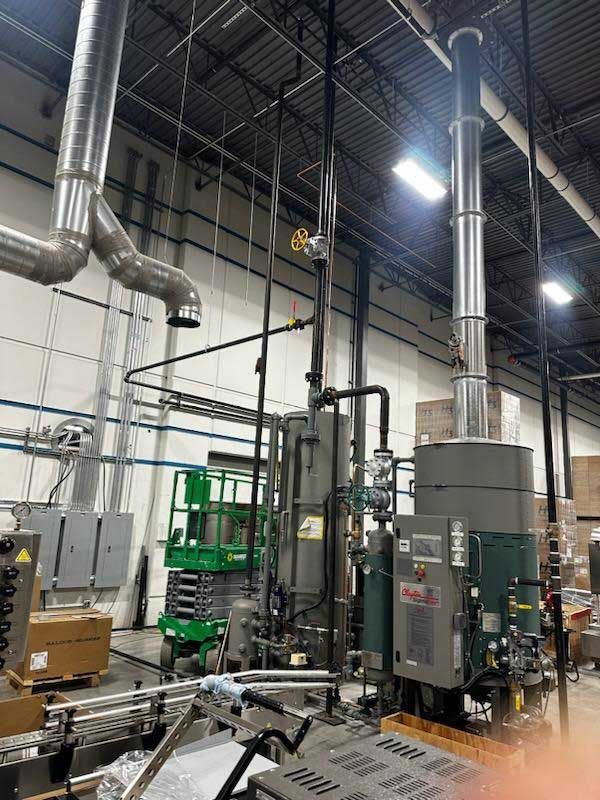 Industrial machinery with pipes and a tall exhaust stack inside a warehouse. Green lift and equipment are visible.