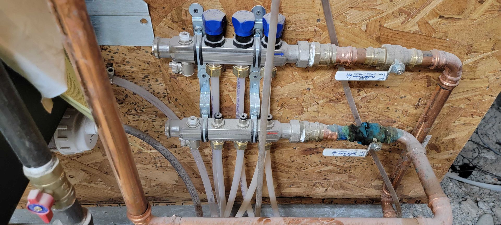 A close-up of a hydronic manifold system with blue-capped valves and clear plastic tubes against a wood panel.