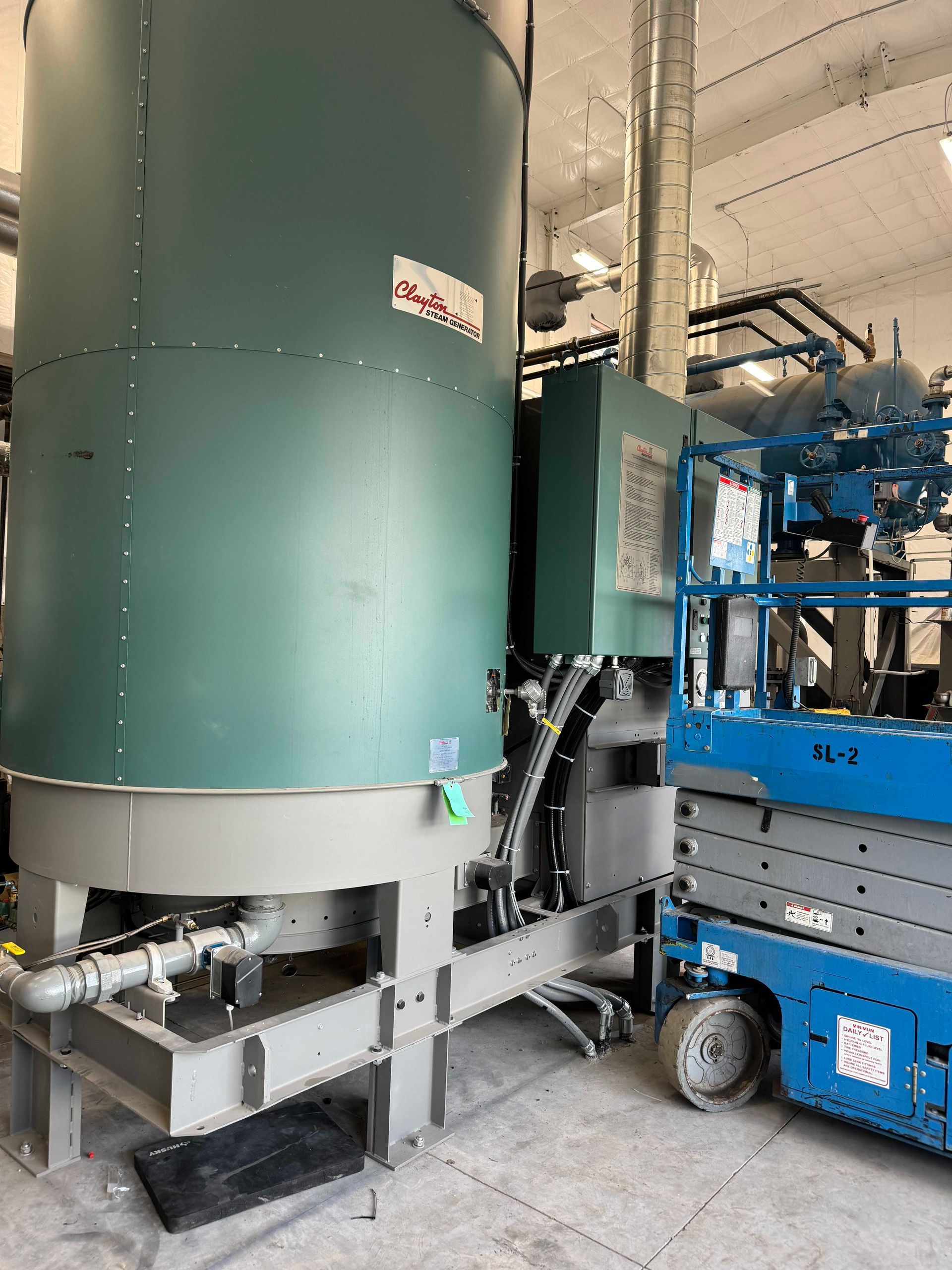 A large industrial machine, green and gray, inside a warehouse. A blue lift is positioned next to the machine.
