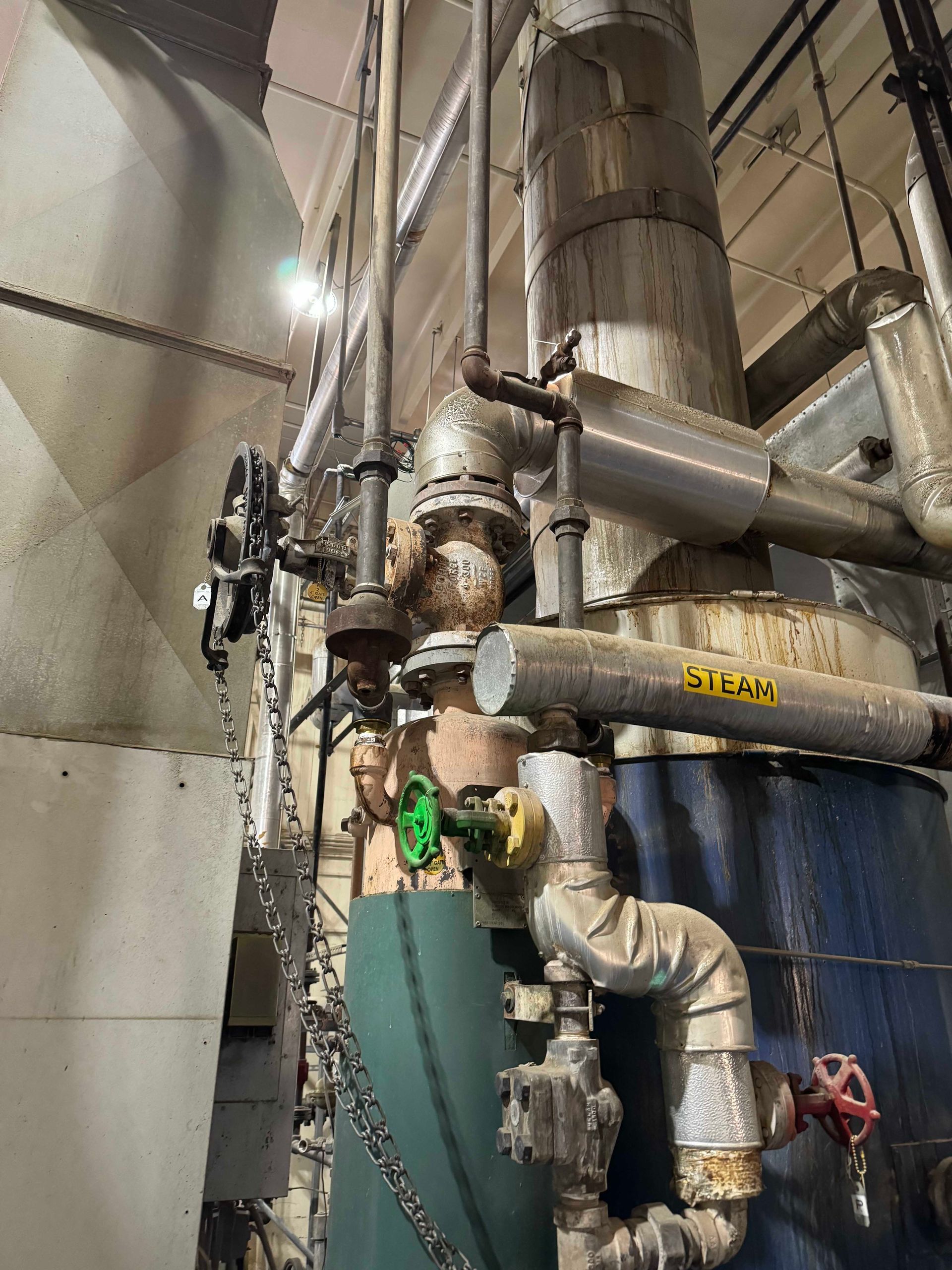 Industrial equipment with pipes, valves, and insulation, in a factory setting. Some pipes have red, green, and yellow handles.