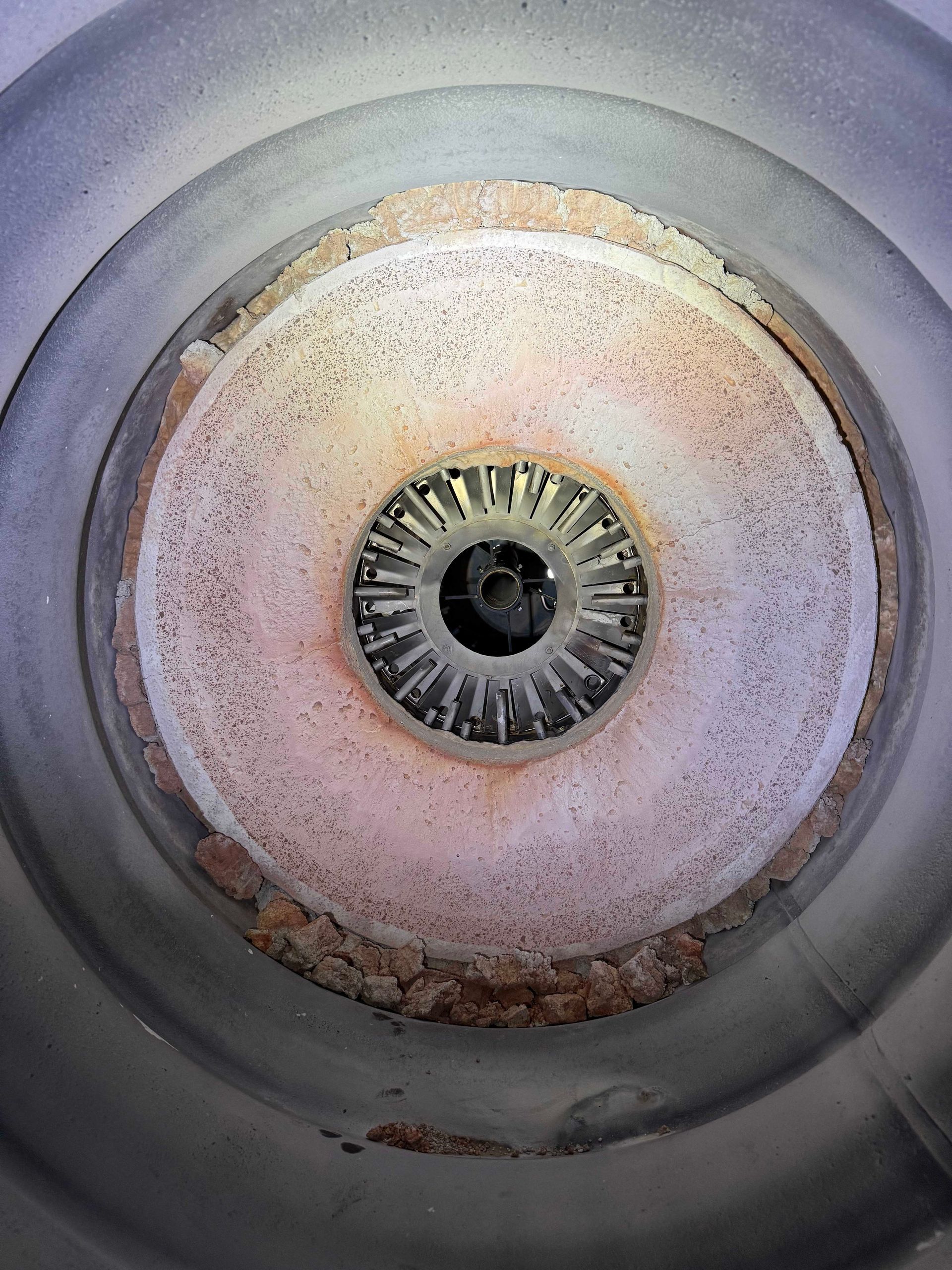 Overhead view of a cylindrical industrial component with concentric rings of light and dark colors, leading to a central dark opening with radial fins.