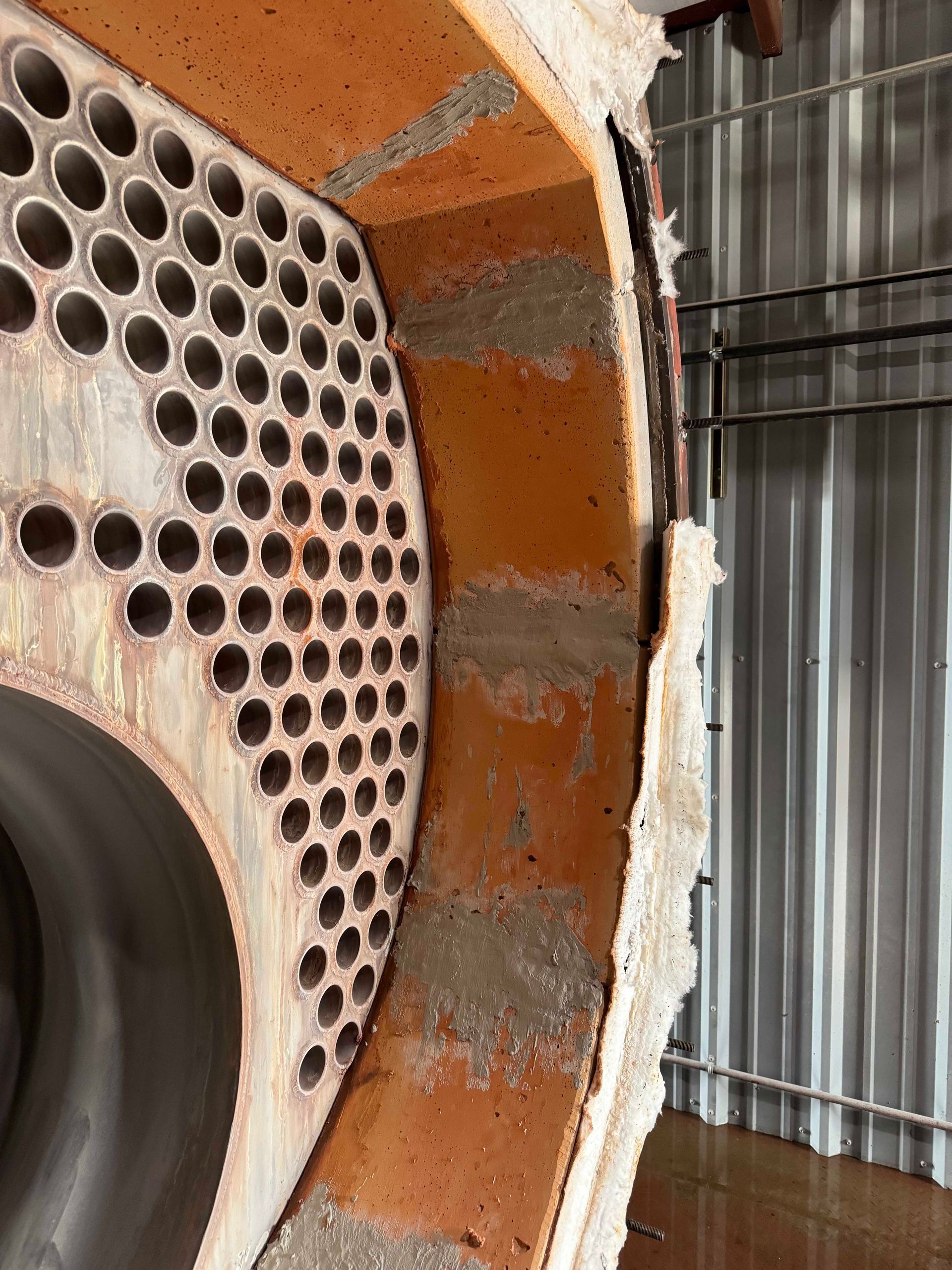 Close-up of a section of a boiler with a metal plate full of circular holes, surrounded by reddish-orange brickwork and white insulation.