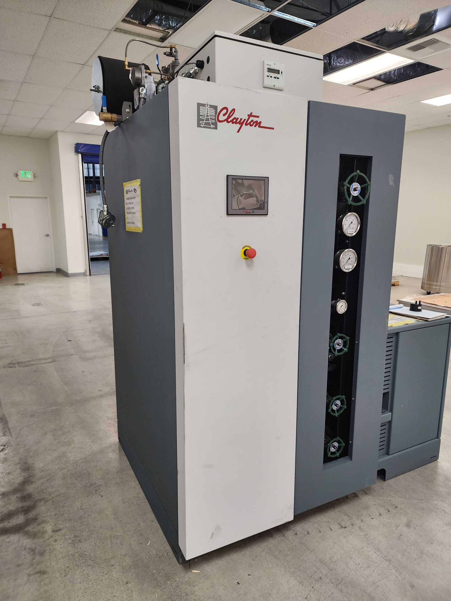A large, industrial printing press with gray and white panels. The press is in a brightly lit room.
