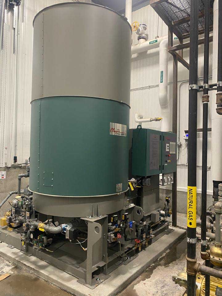 A large, cylindrical industrial water heater with teal and gray casing sits on a concrete platform inside a white-walled room.