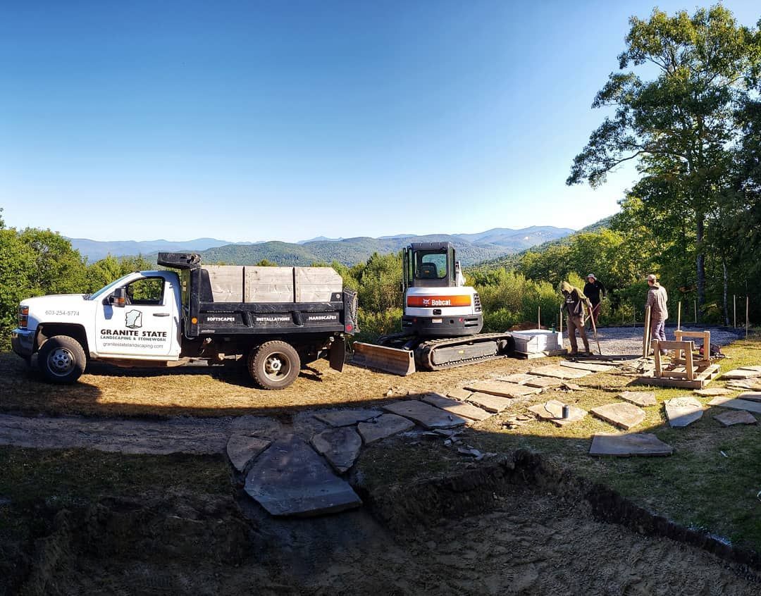 Workers operate a small excavator and dump truck on a site with stone pavers amidst a mountainous outdoor landscape.