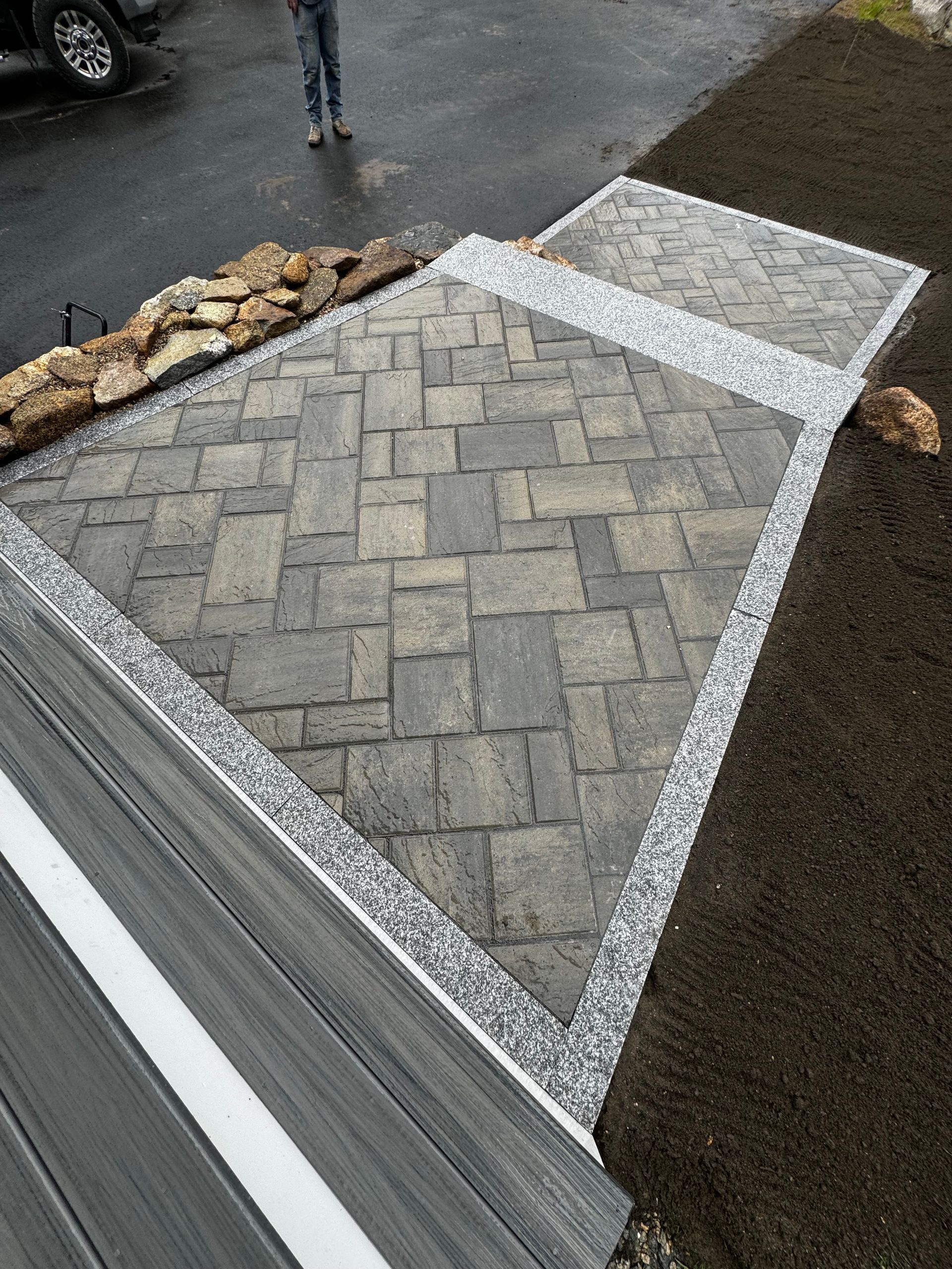 A stone paver walkway with a herringbone pattern, framed by gravel, leading from a dark deck to a landscaped yard.