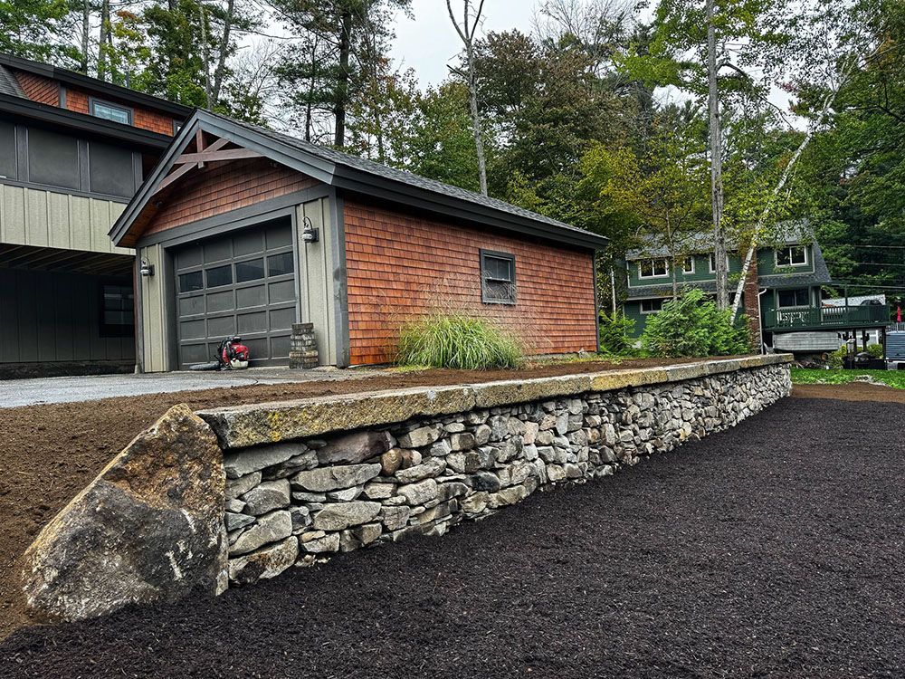 A stone retaining wall sits in front of a brick garage with a dark shingled roof, surrounded by mulch and trees.