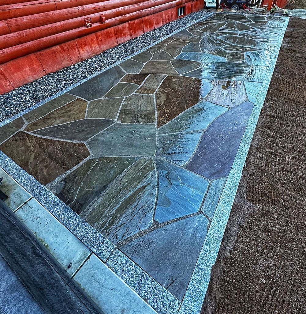 A stone patio walkway with irregular, multicolored flagstones bordered by granite pavers next to a red log building.