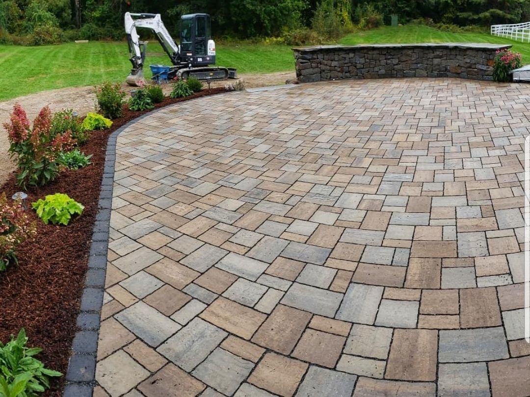 A newly installed paver patio in a backyard with a curved stone retaining wall and a small excavator in the background.