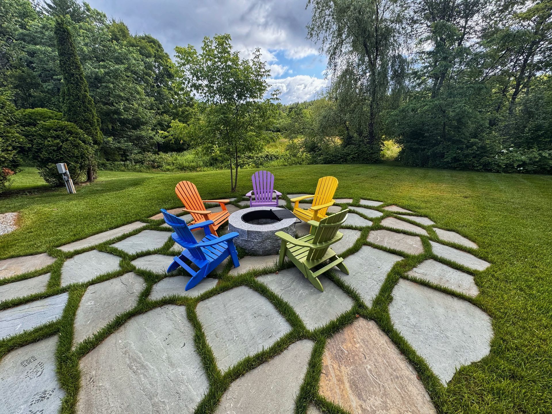 A stone patio with a fire pit and five colorful Adirondack chairs arranged in a circle on a green lawn.