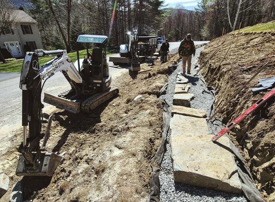 A mini excavator sits on a dirt road next to workers building a stone wall along a gravel-lined hillside trench.