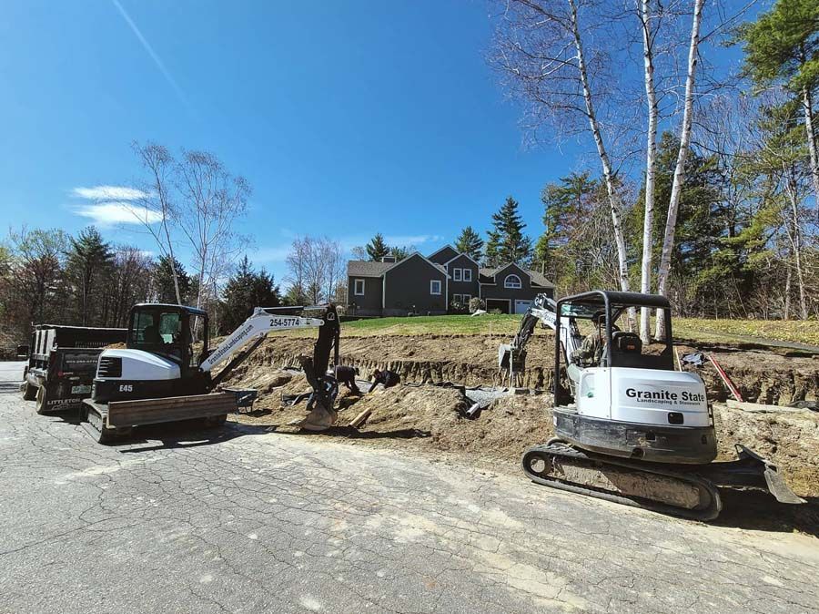 Two white excavators on a gravel driveway in front of a residential house during a daytime landscaping project.