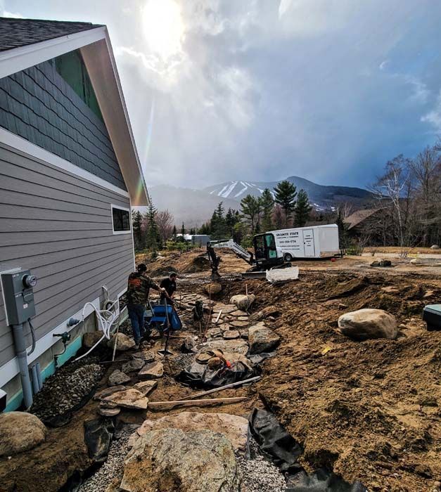 Construction workers install a stone walkway next to a grey house with mountains in the background under a cloudy sky.