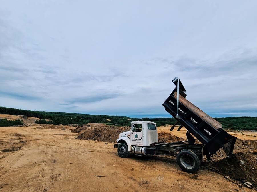 A white dump truck sits on a dirt lot with its black cargo bed raised, dumping a load of soil onto the ground.