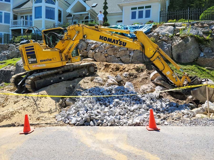 A yellow Komatsu excavator sits on a dirt slope near a residential home, with a pile of rocks and orange traffic cones.