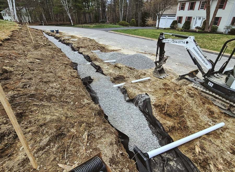 A long, gravel-filled trench with drainage pipes being installed in a residential yard near a house and small excavator.