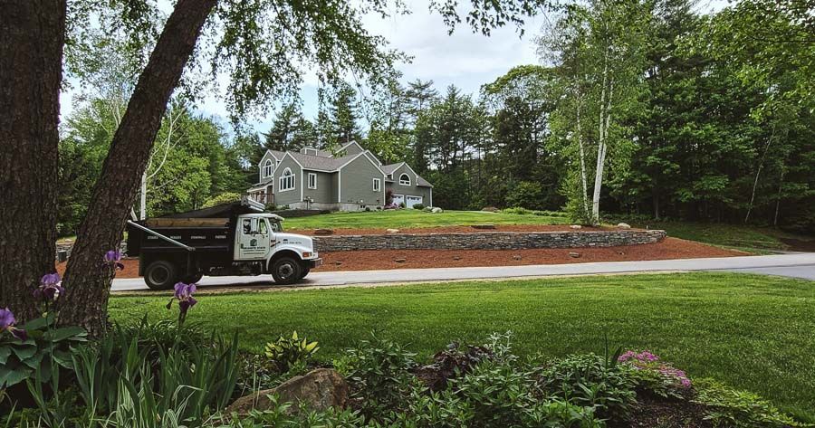 A dump truck sits on a driveway in front of a gray house with a stone retaining wall and a lush, green yard.