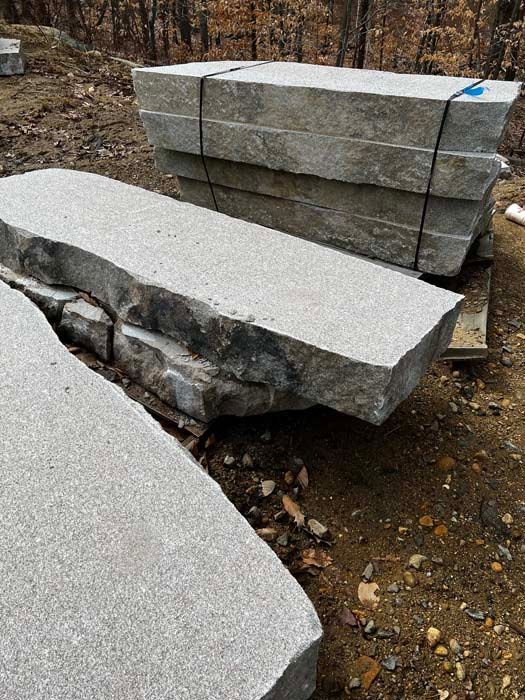 Large, light-gray granite stone slabs stacked on a pallet outdoors on a dirt ground with trees in the background.